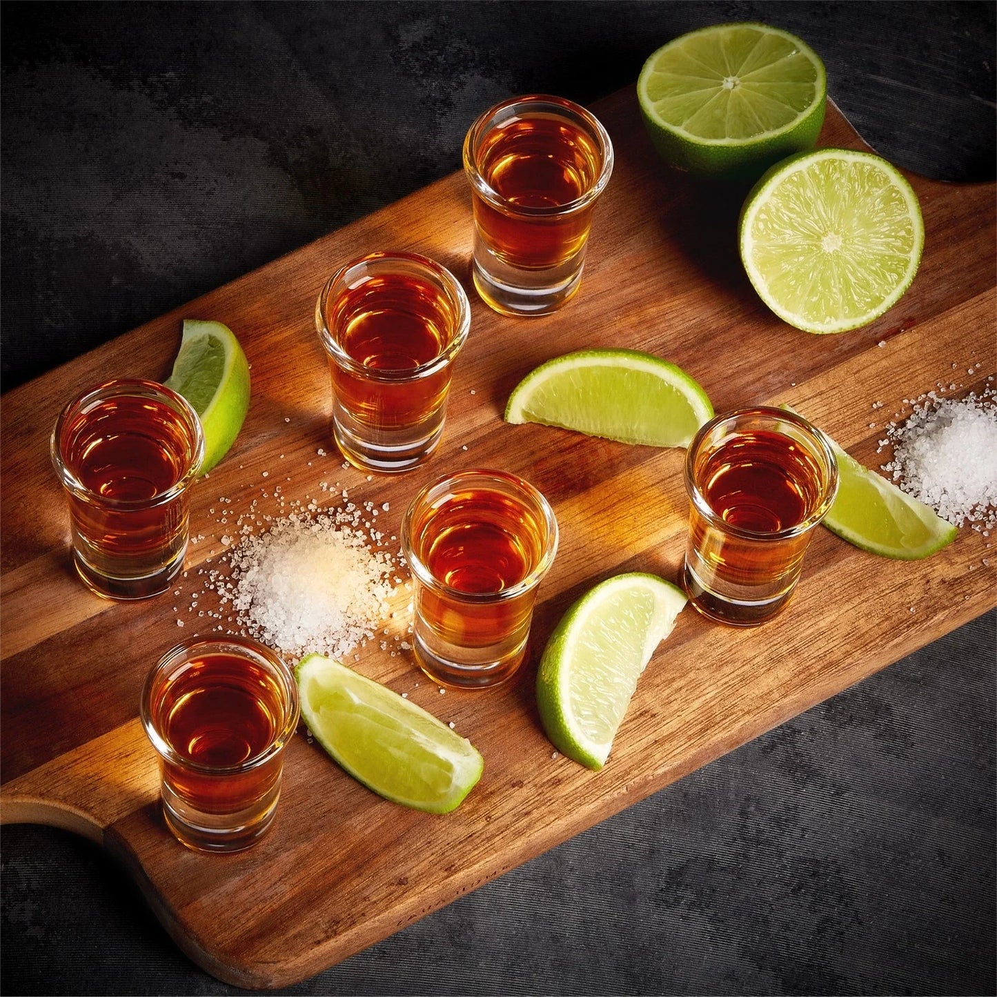 Set of shot glasses with amber liquid on a wooden board with lime wedges and salt.